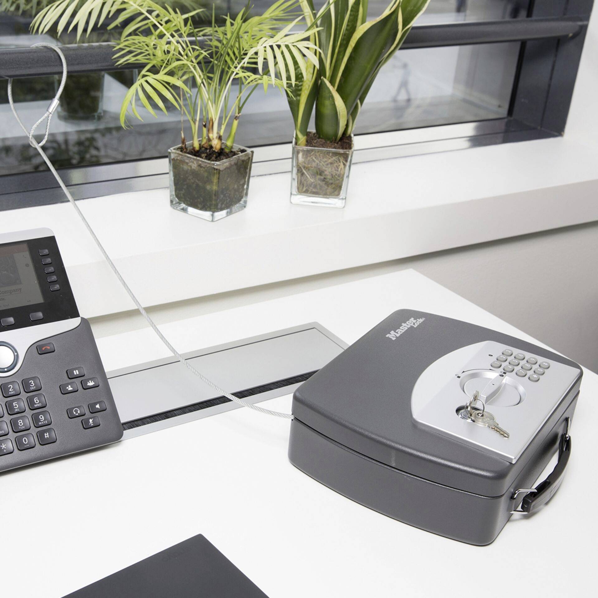 'Office desk with telephone and small security safe, locked with a bunch of keys on top. Two plants in the background.'