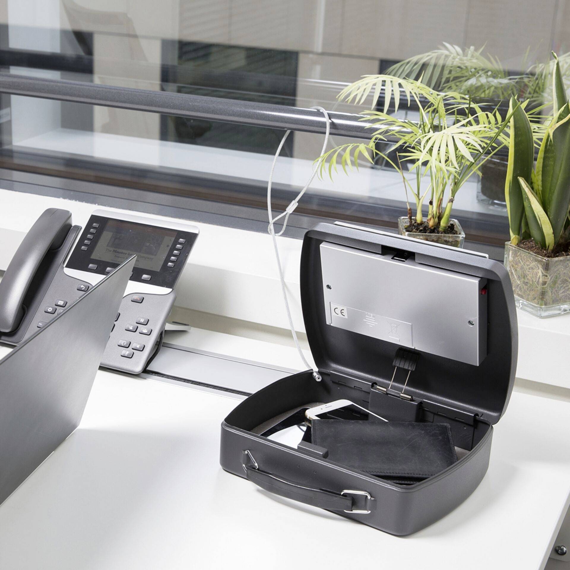 An open safe on a desk near a window, with a telephone beside it and a small plant nearby.
