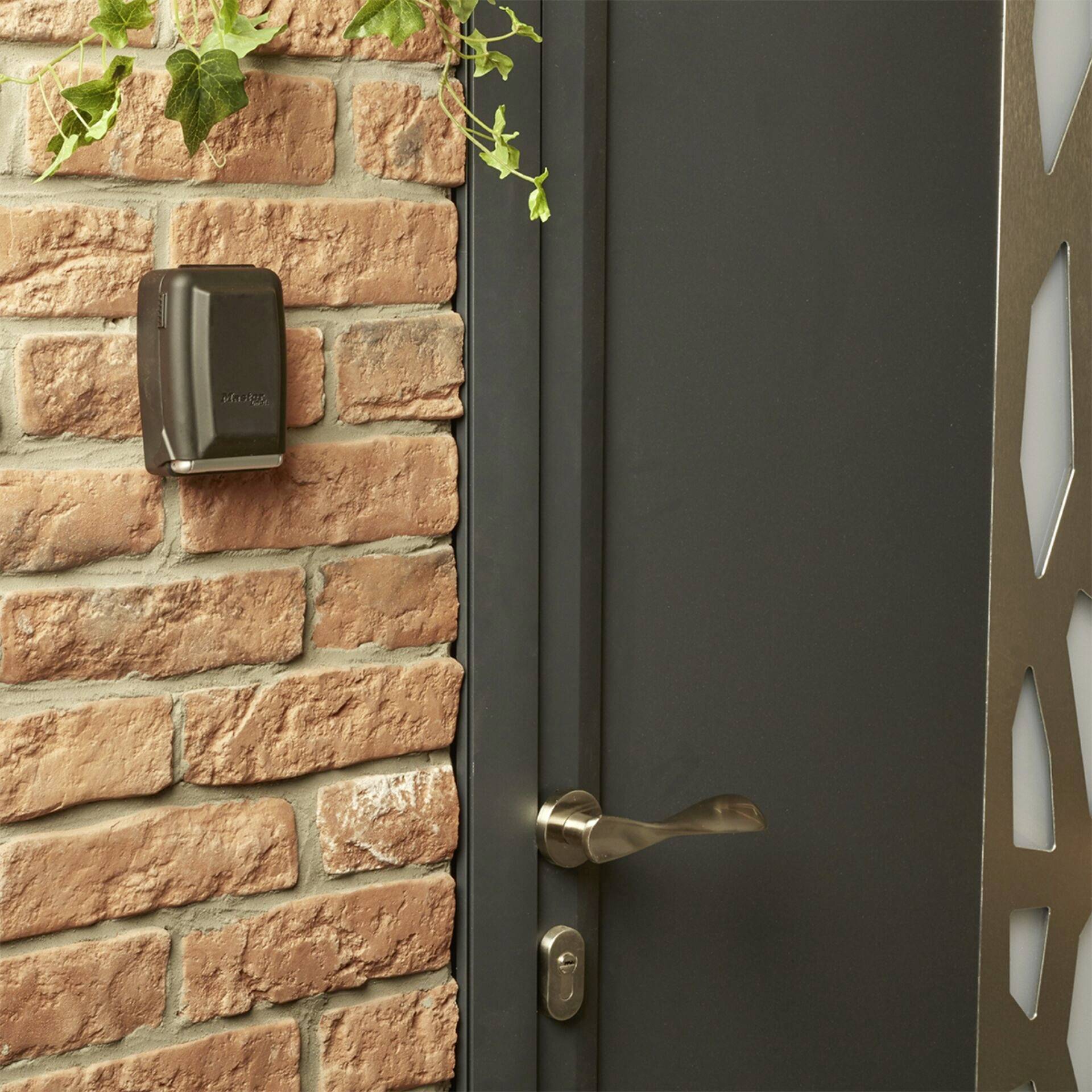 The close-up shows a grey door with a modern door handle, next to a red brick wall with an access control device and hanging plants.