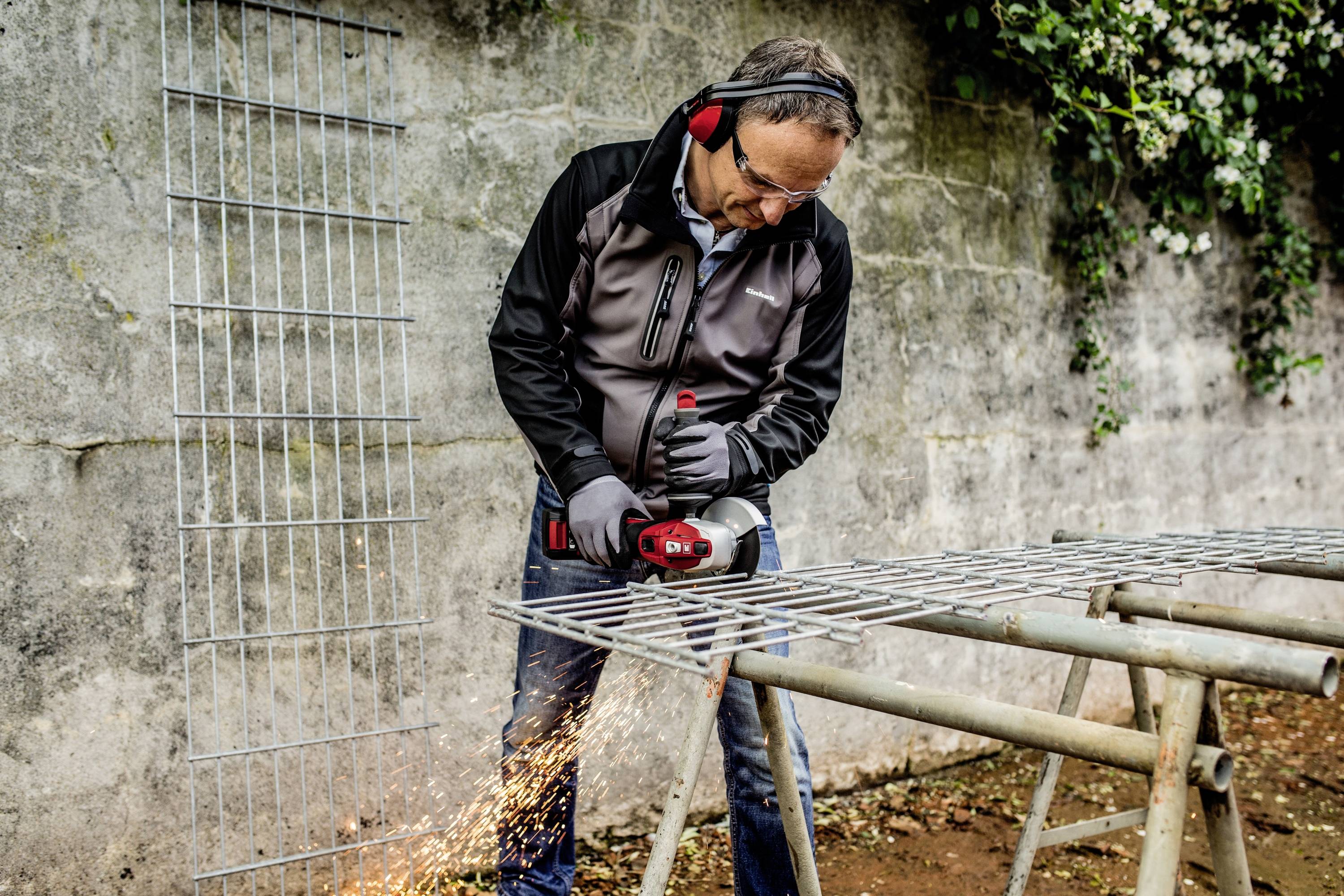 A man wearing ear defenders and safety glasses is cutting a large metal grid with an angle grinder. Sparks are flying.