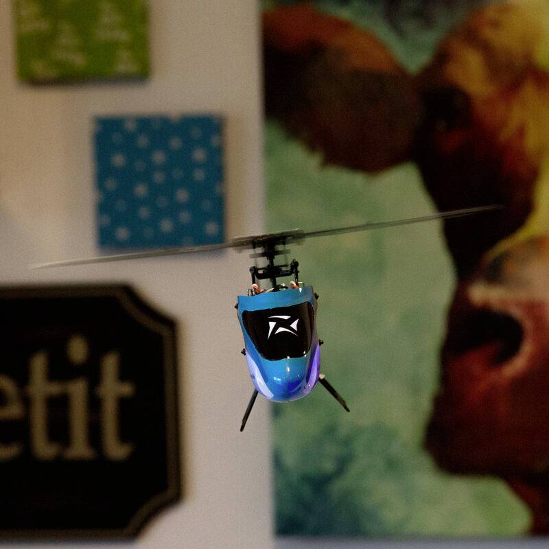 A small blue model helicopter hovers in a room, surrounded by blurred wall decorations in the background.