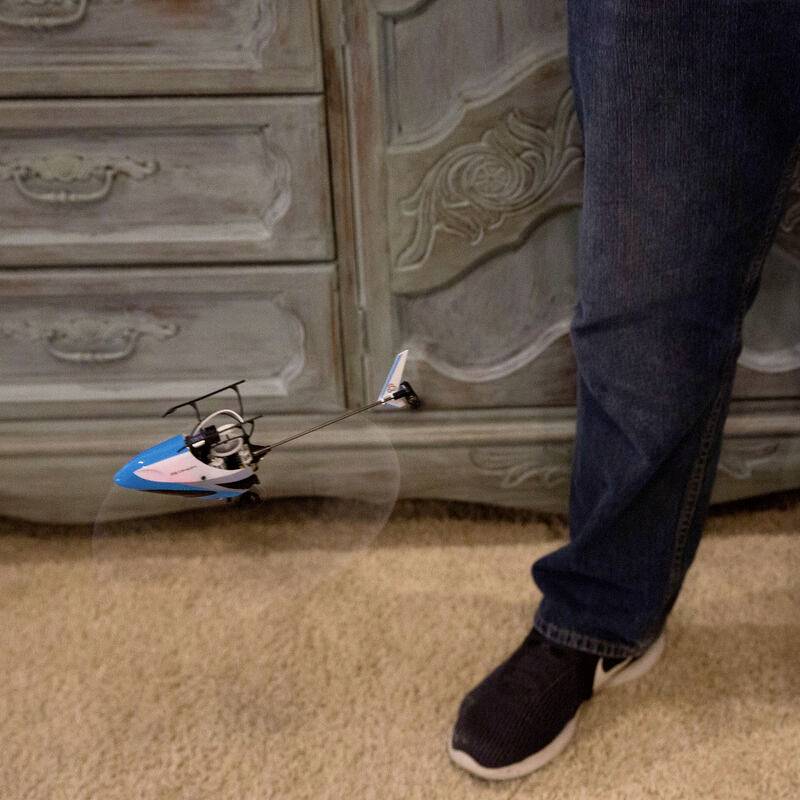 A person is standing on a carpet next to a small blue remote-controlled helicopter that is flying.