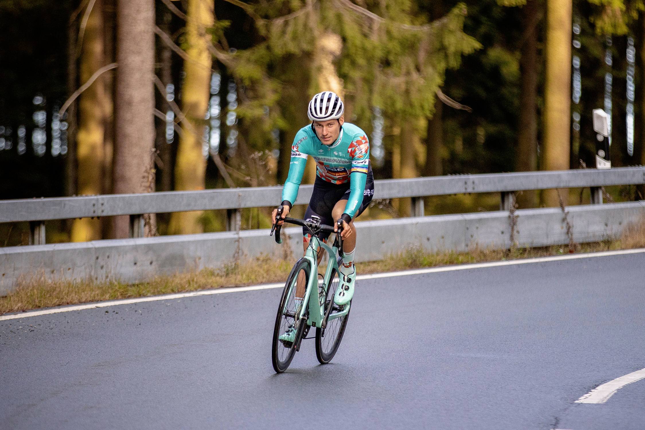 A cyclist in turquoise clothing is riding along a winding road through a wooded area.