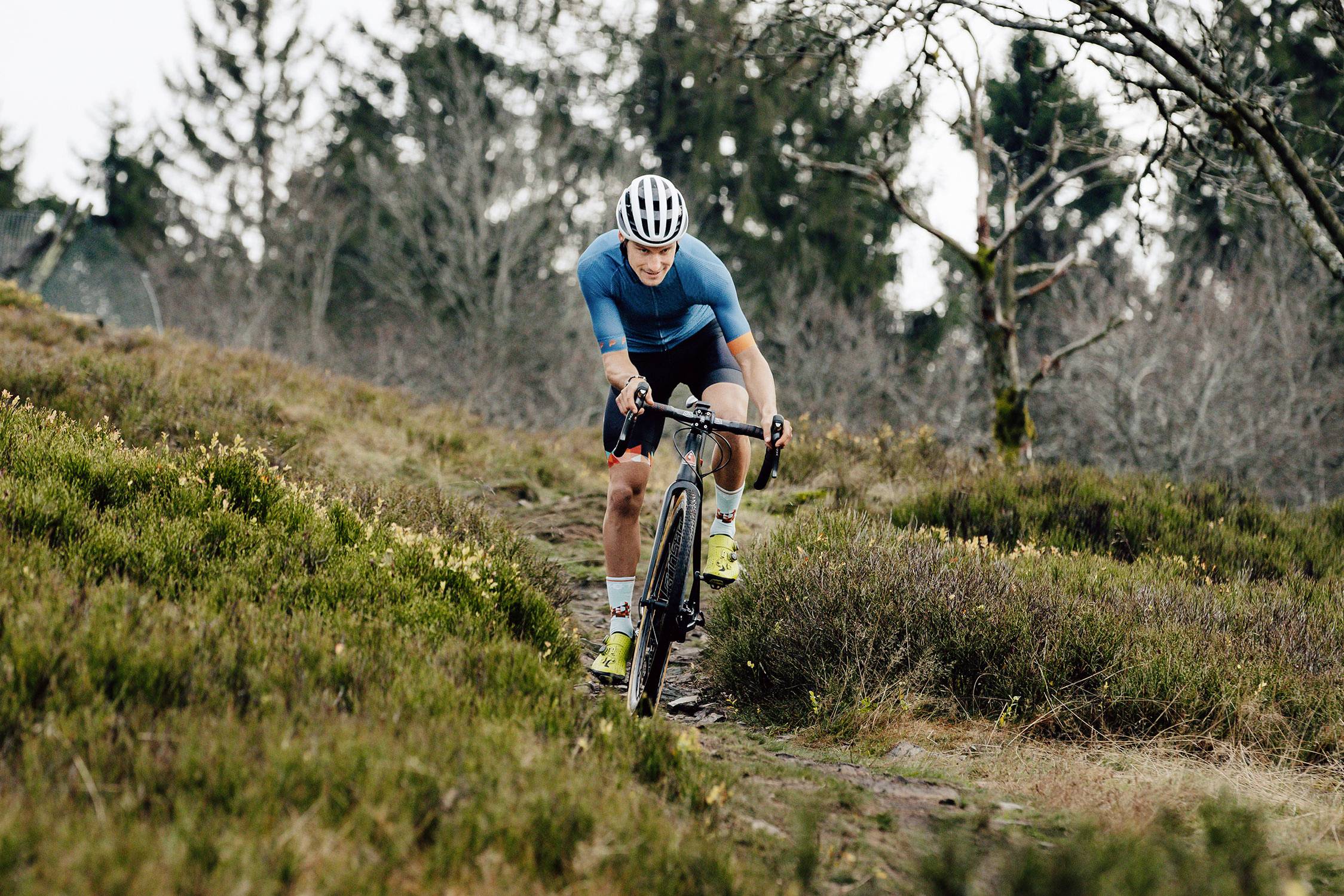 A person is cycling along a narrow, natural path through a wooded landscape. They are wearing a helmet and sporting attire.
