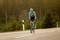 A cyclist in sports attire is riding along an empty country road through a wooded landscape.