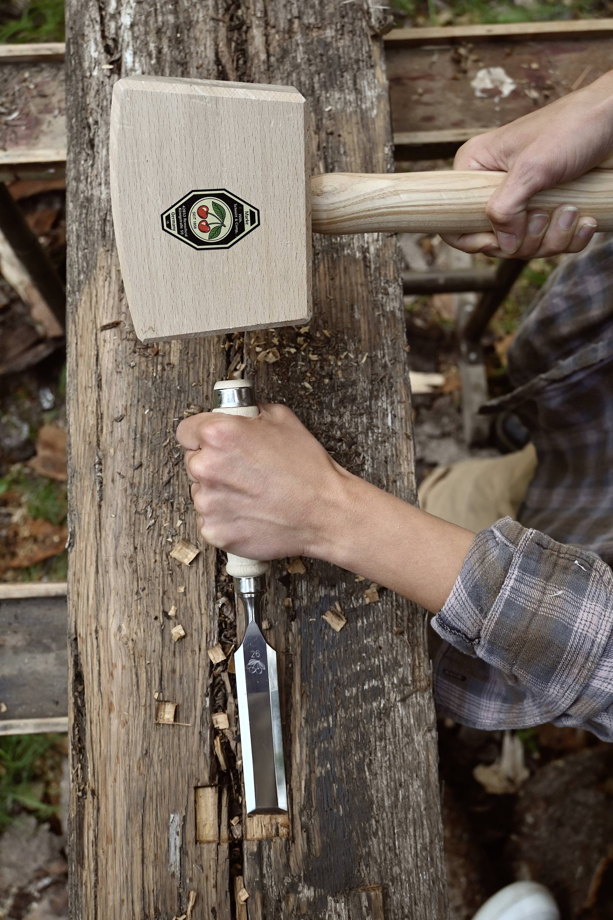 A person is working on a piece of wood with a hammer and chisel, possibly to create an indentation. The focus is on the tools.