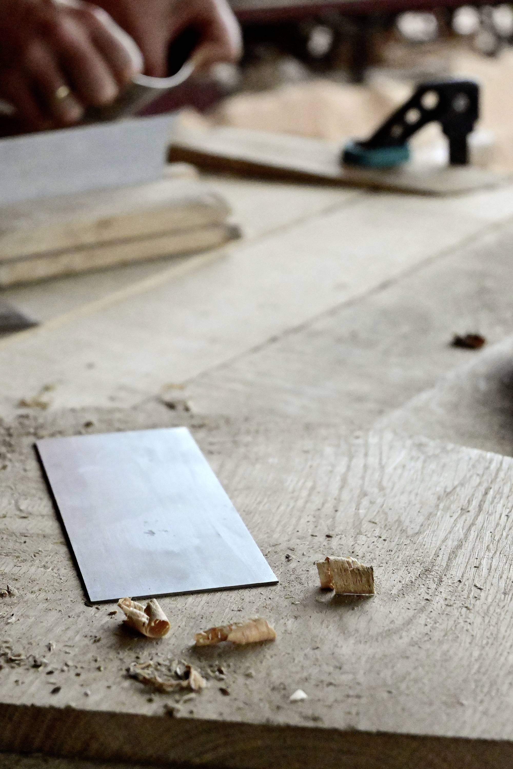 A woodworker planes wood on a workbench. Wood shavings are scattered, and a metal tool lies in the foreground.