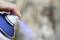 A hand presses the nozzle of a blue spray can, with a fine spray mist emerging. Background blurred.