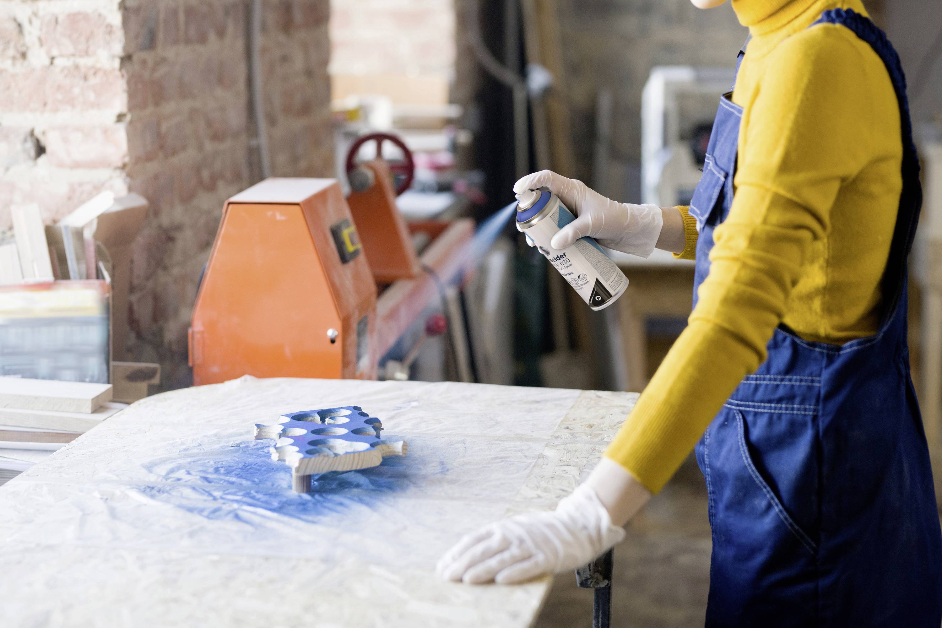 A person in a yellow turtleneck jumper and blue dungarees is spraying blue paint onto a wooden workpiece in a workshop.