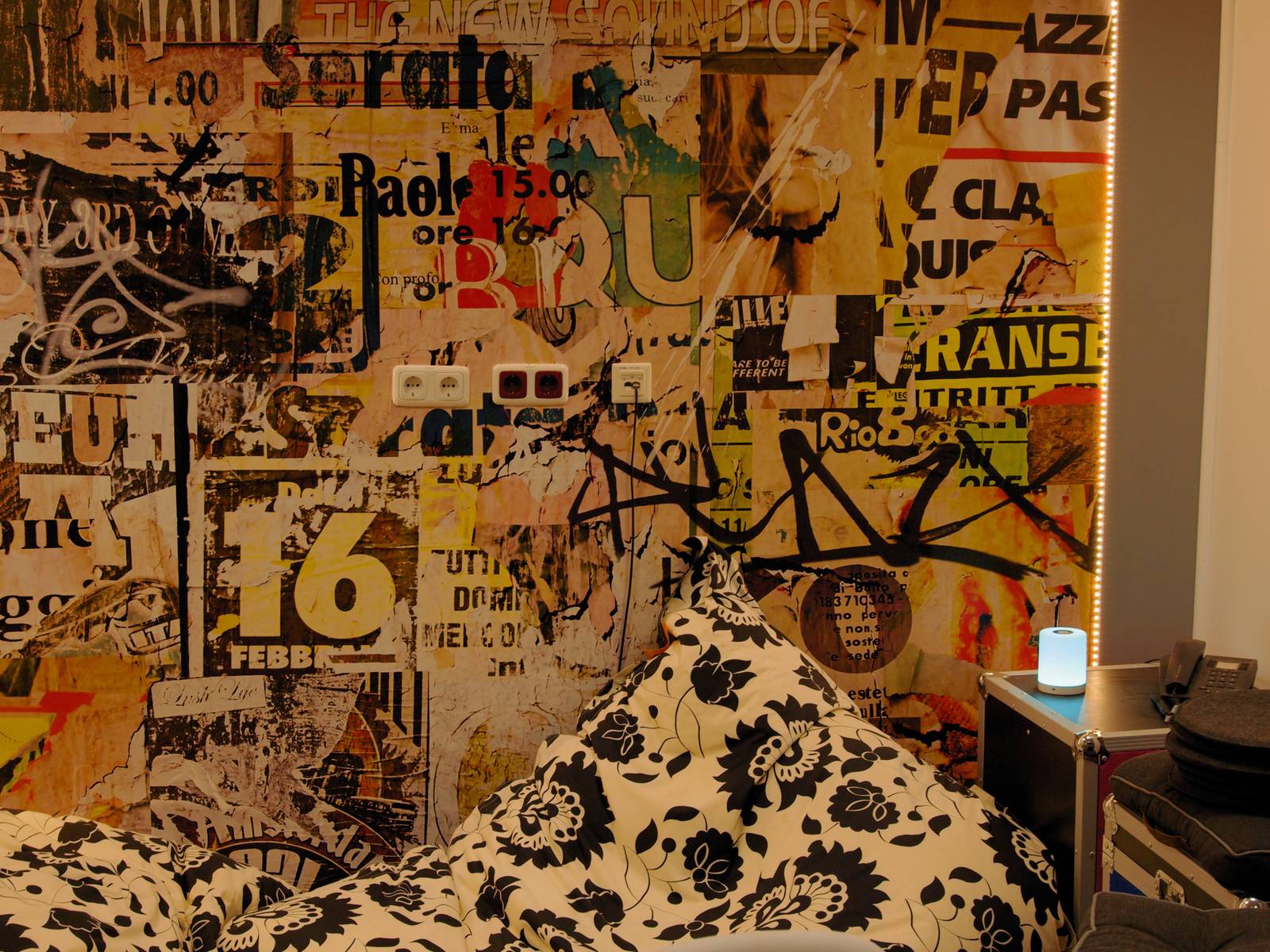 Wall with colourful, overlapping posters and graffiti. In the foreground is a bed with patterned cushions and a small blue lamp.