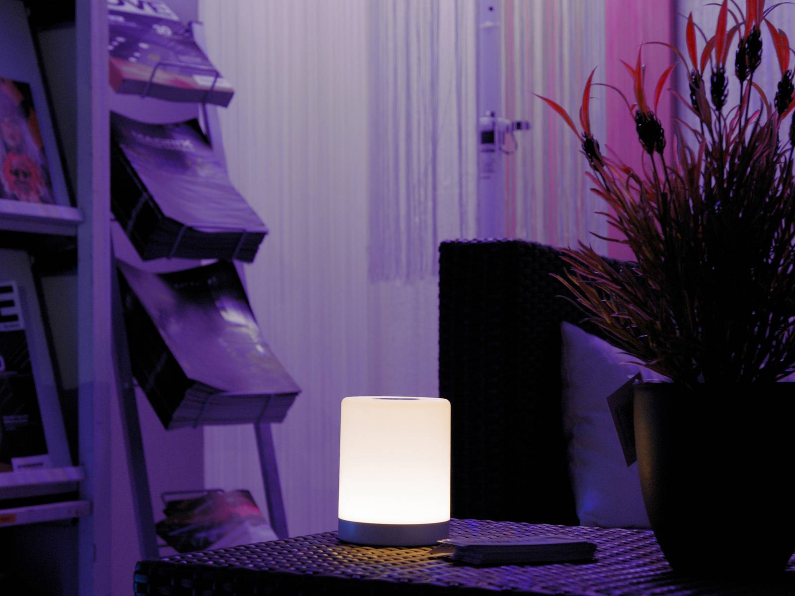 A glowing table lamp on a dark table in a room with a magazine rack and plant in the background.