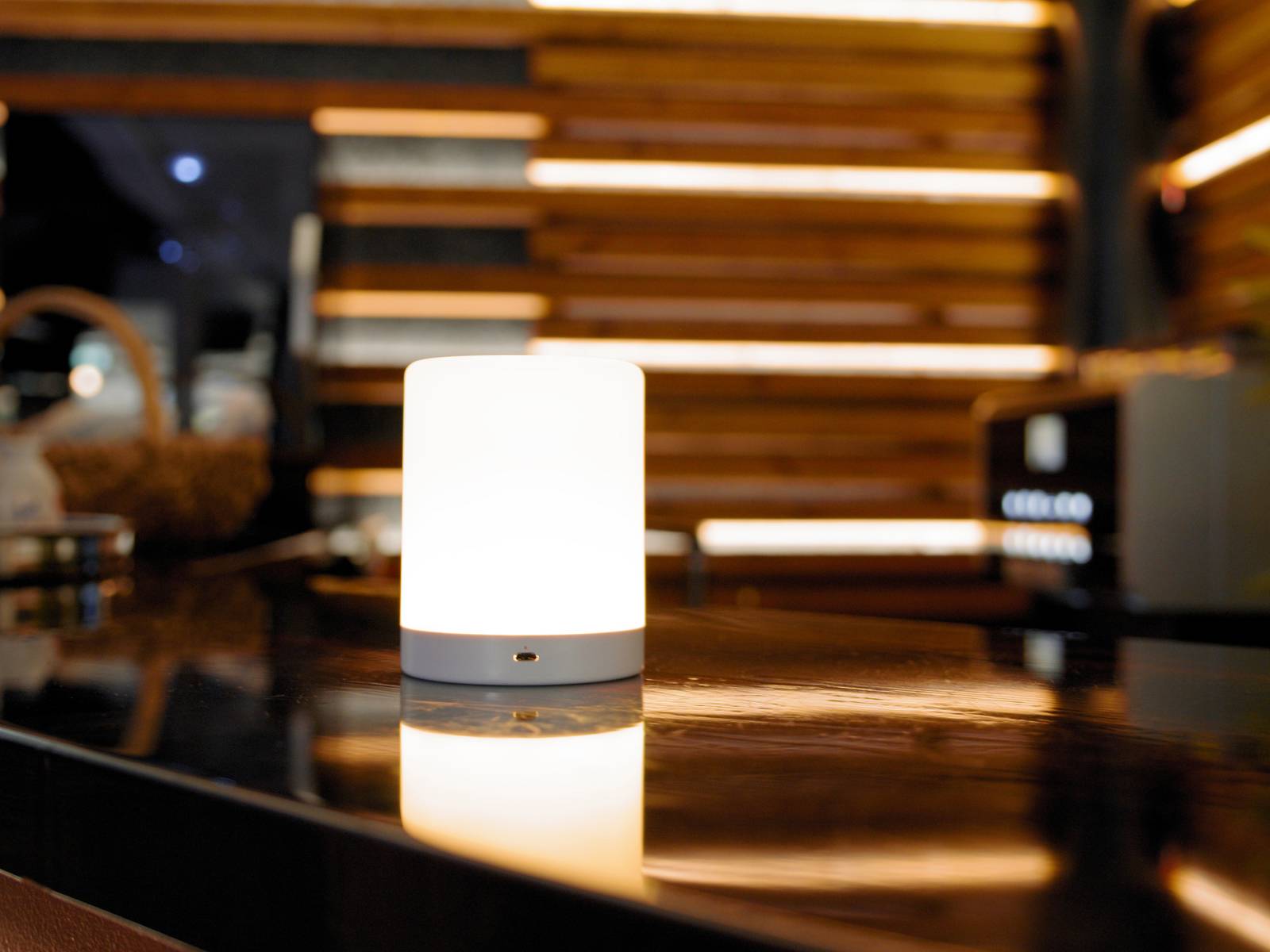 A table lamp with soft, warm light sits on a dark, glossy table in front of a wooden panelled background.