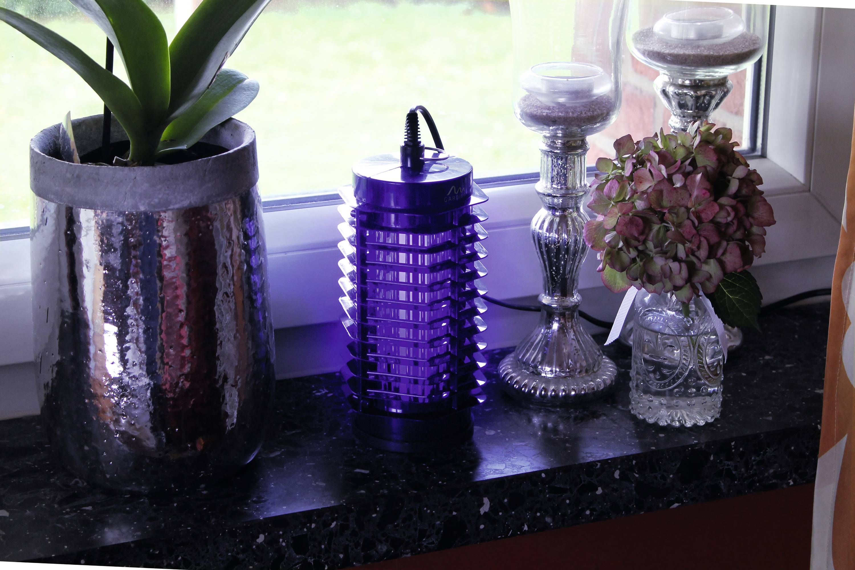 A windowsill with a plant, a purple insect killer, and two candlesticks with flowers.