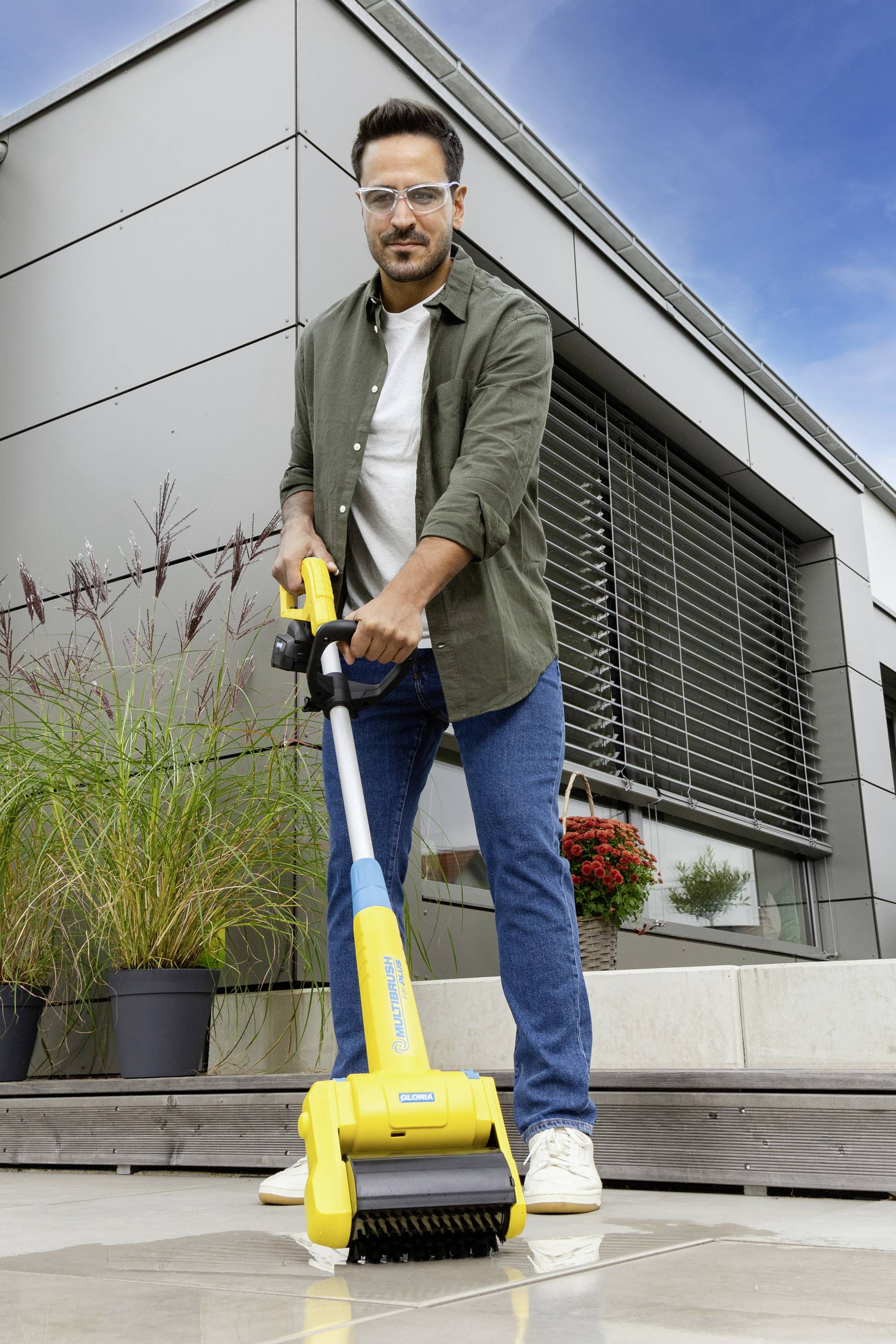 A man is cleaning a terrace with a yellow floor cleaner. Behind him is a modern building with large windows and plants.