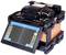 A splice machine with a digital display shows operating modes and settings. The device has a robust construction and functions for optical fibre connection.