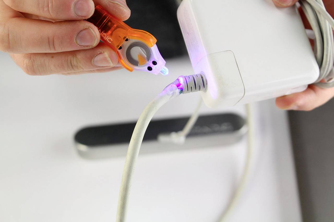 A person is checking a white charger for electrical voltage using an orange device while it is plugged in.