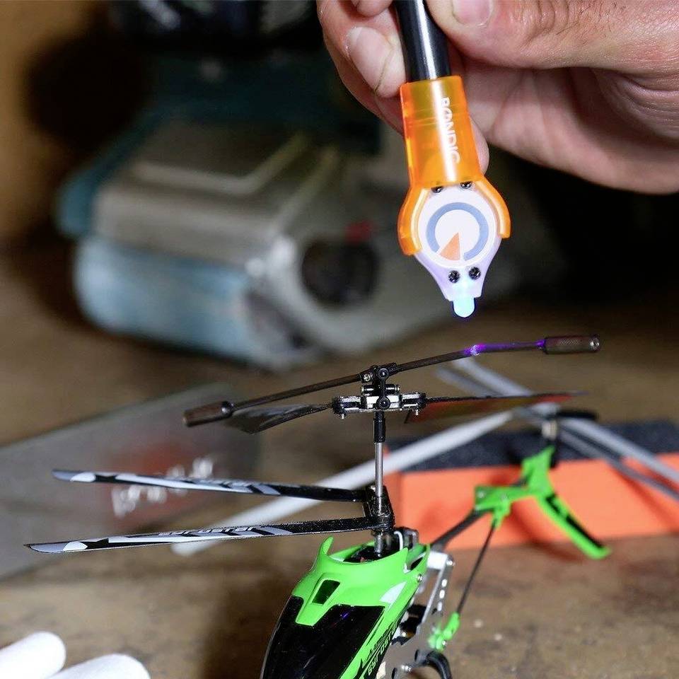 A hand holds a tool over a model helicopter rotor blade. The focus is on the precise adjustment of the rotor blades.