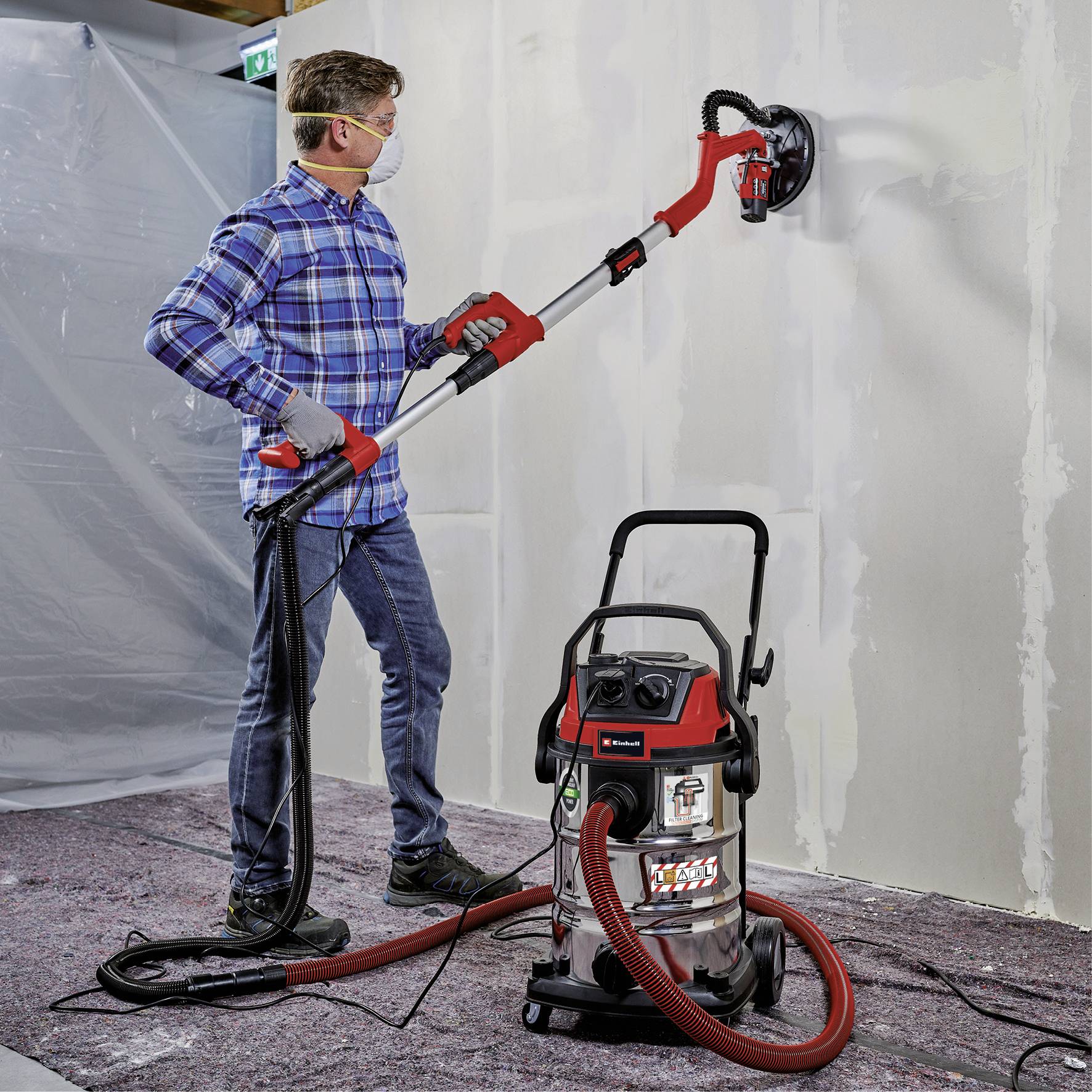 A man is sanding a wall with a large electric sander while wearing a protective mask. A vacuum cleaner is connected to collect dust.