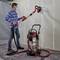 A person is sanding a plasterboard wall using an electric wall and ceiling sander. An industrial vacuum cleaner is collecting dust.