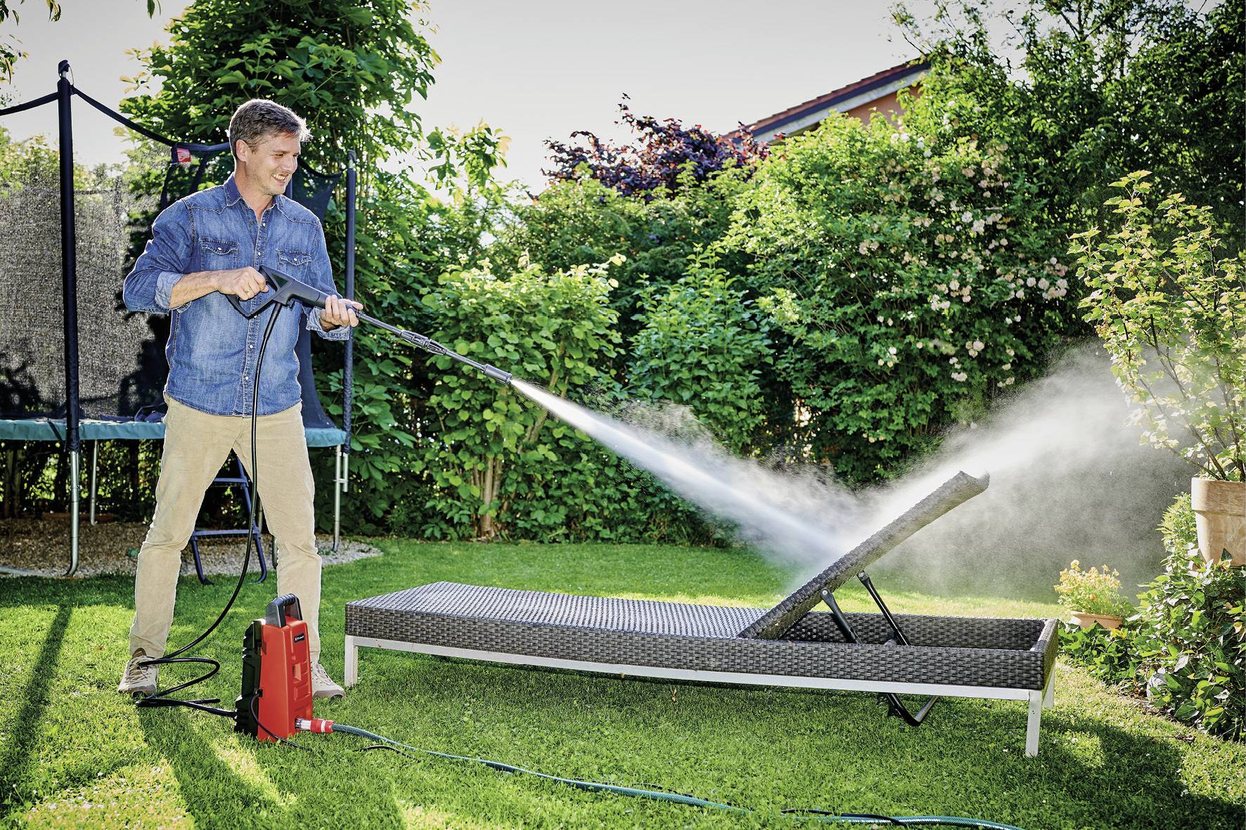 A man is cleaning a garden lounger with a pressure washer in a sunny garden. Shrubs and a trampoline can be seen in the background.