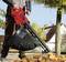 A person is using a red leaf blower on a pavement. The leaf blower is sucking leaves into a black bag. Trees and a house are visible in the background.