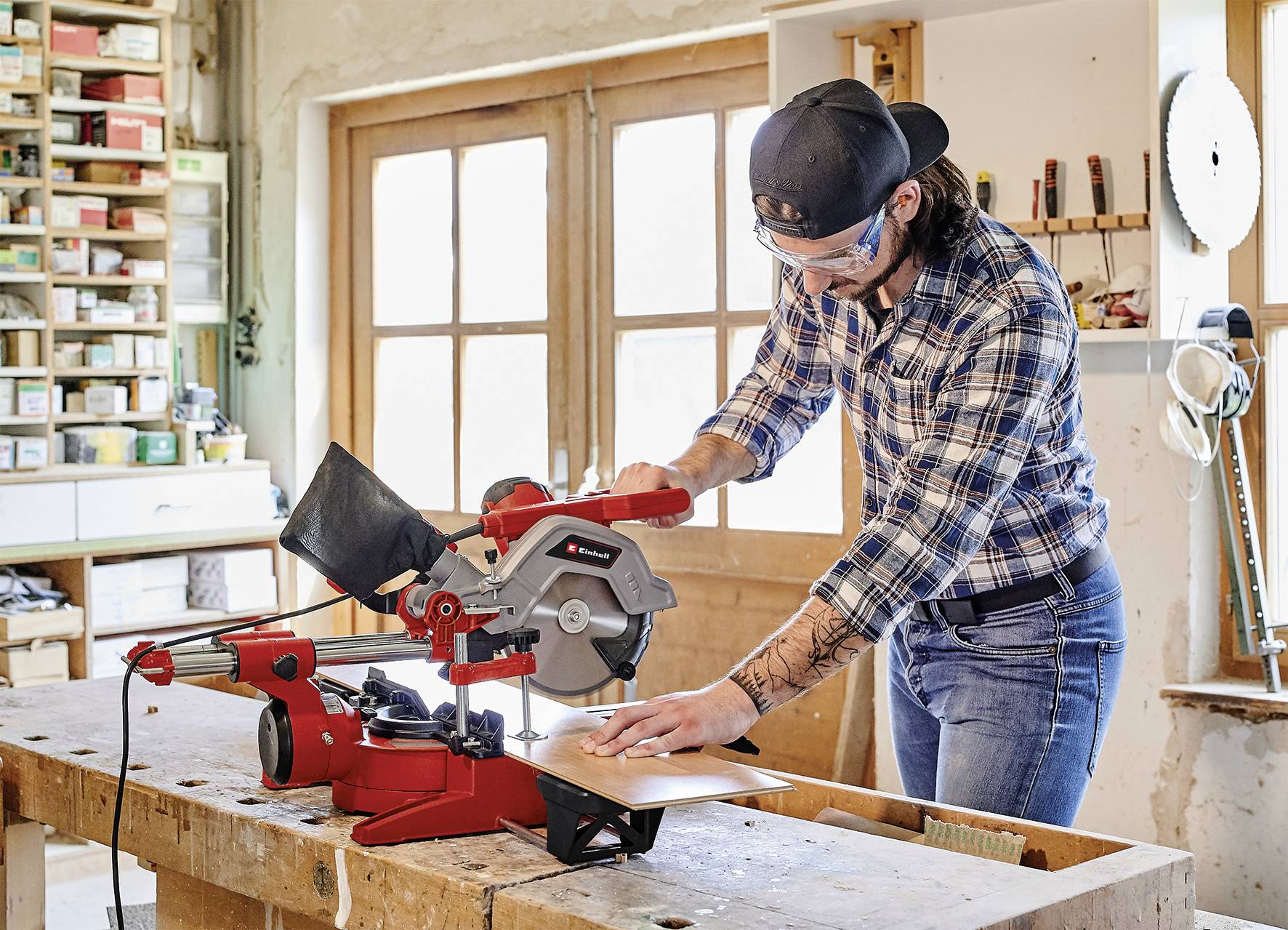 A man wearing safety glasses and a cap is cutting a wooden board with a circular saw on a workbench in a workshop.