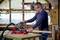 A man is working in a workshop with a red circular saw, cutting a piece of wood. Tools and shelves are visible in the background.