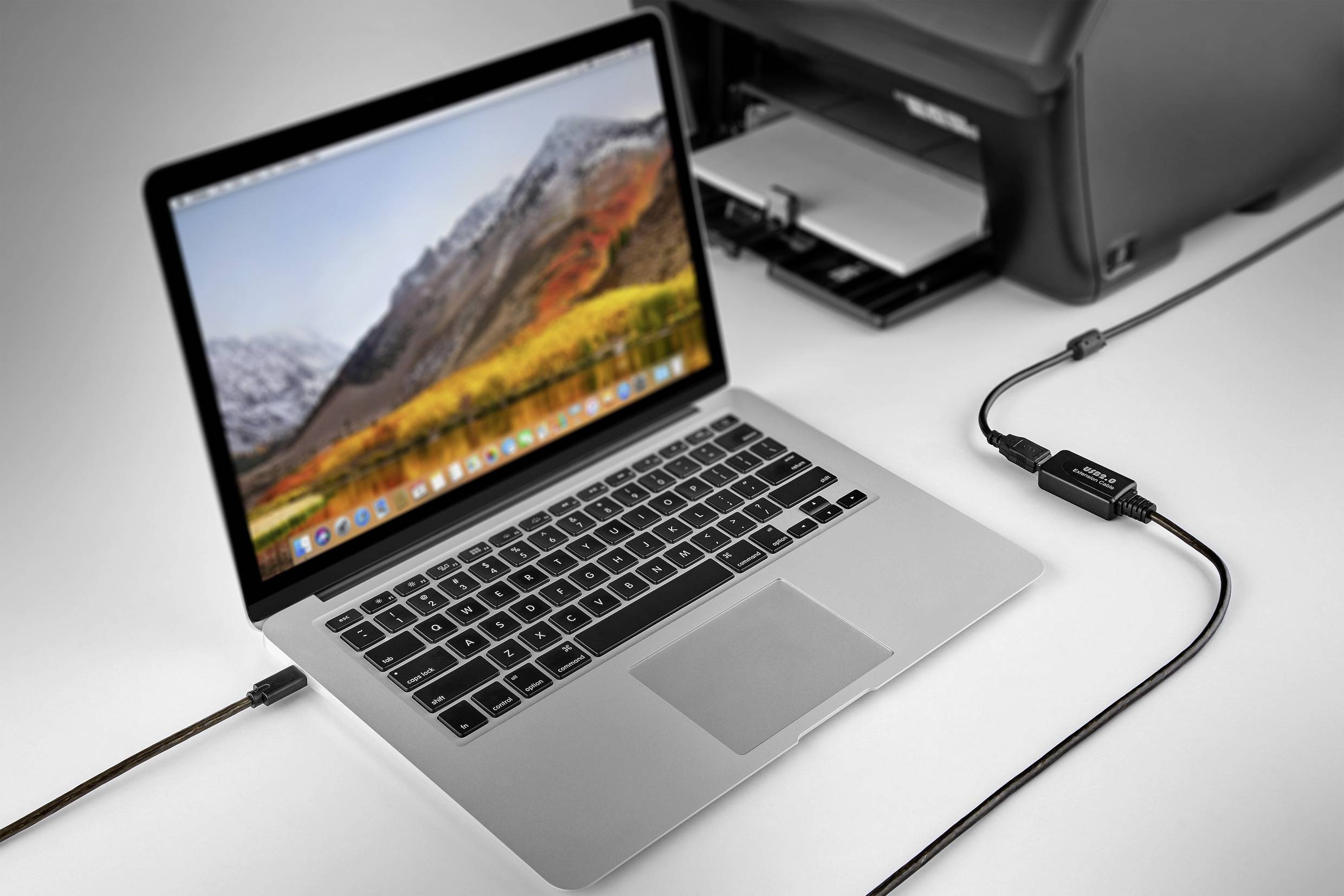 A laptop is connected to a printer via a cable. The laptop displays a mountain landscape on the screen.