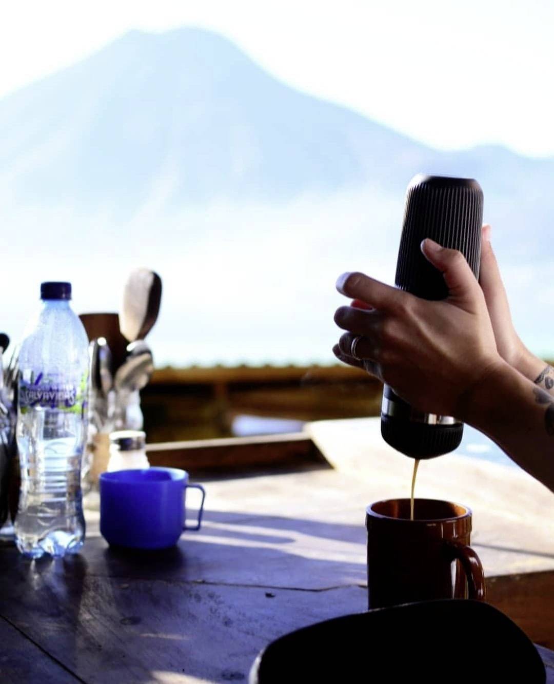 A person is preparing coffee using a hand device, with a mountain visible in the background during daylight. On a table, there are mugs and a water bottle.