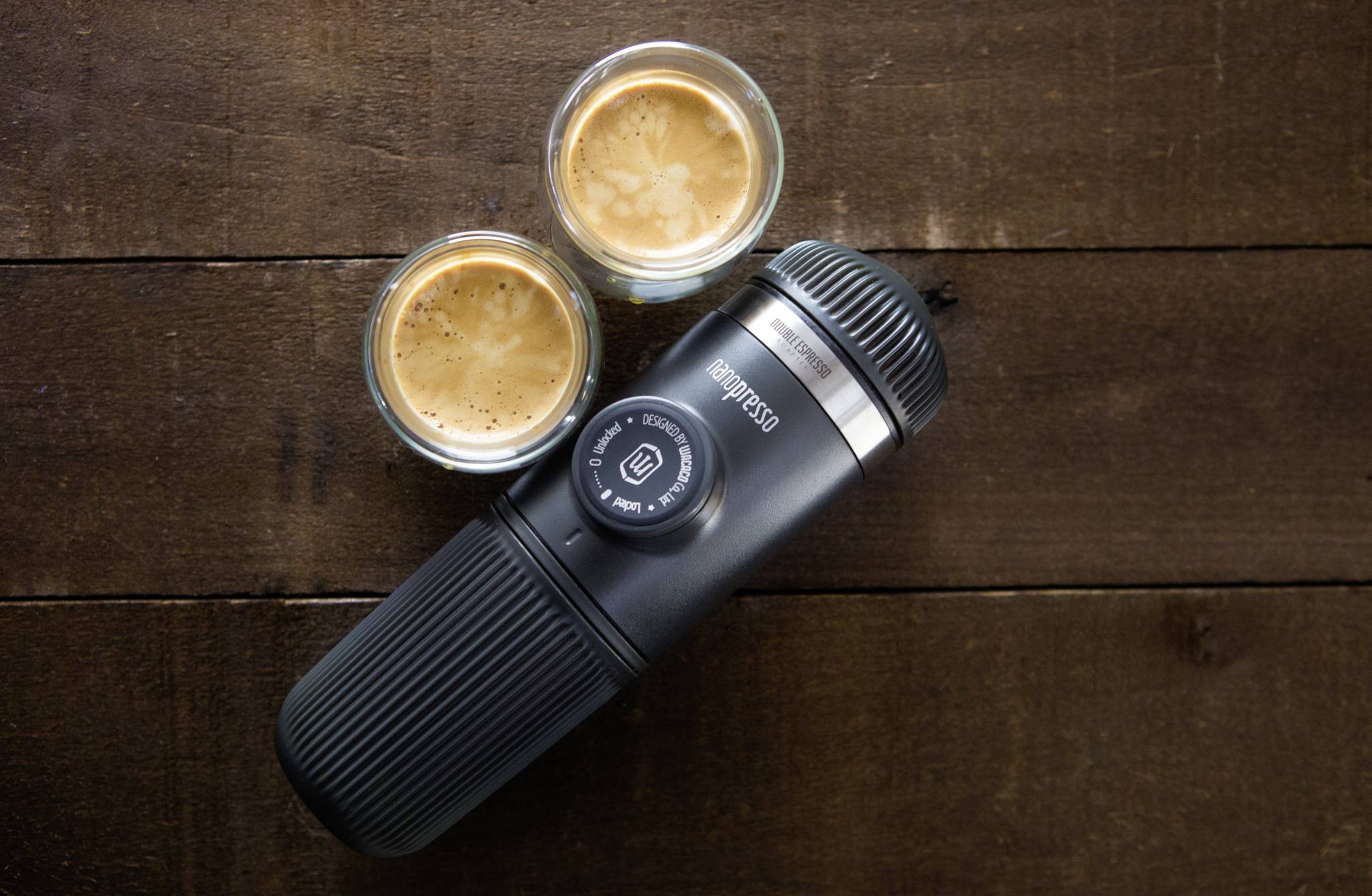 Portable coffee machine and two espresso glasses on a wooden table; the machine is black and cylindrical, glasses with a foamy top.