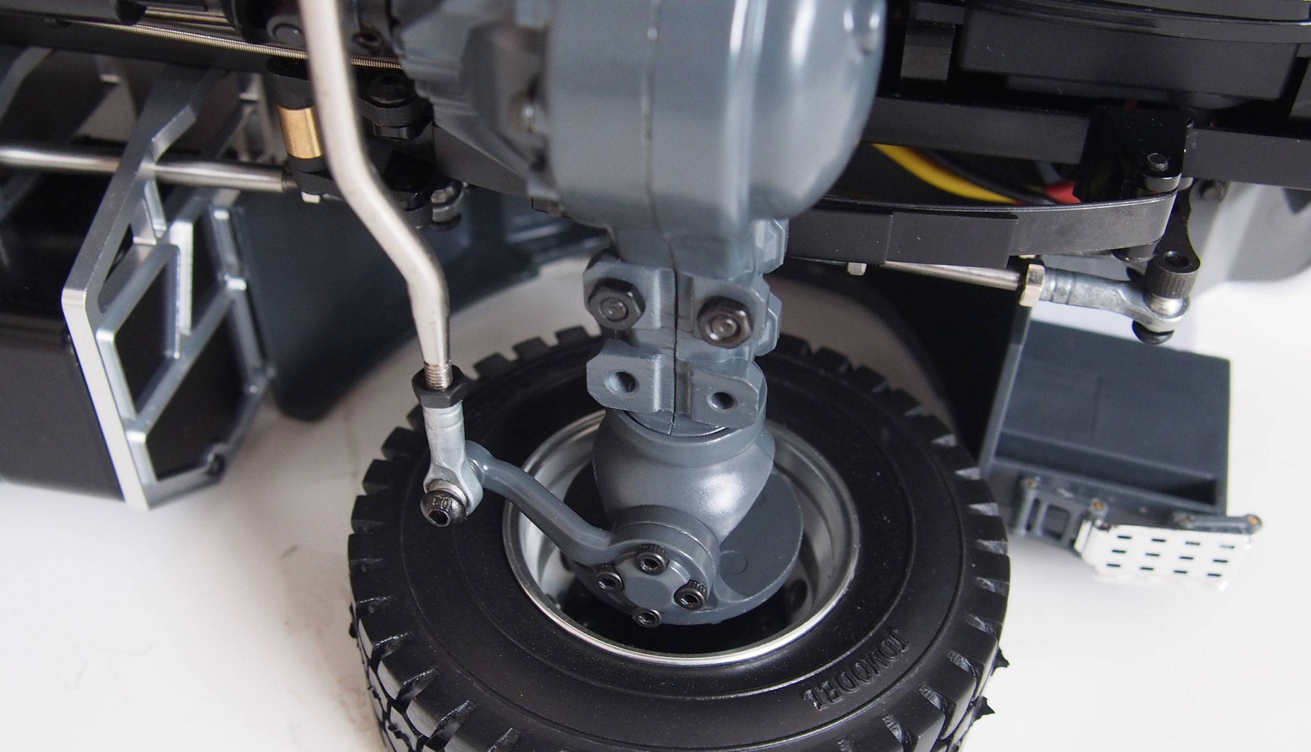 Close-up of a model vehicle chassis focusing on a wheel and parts of the suspension. Tyres, steering, and suspension are clearly visible.