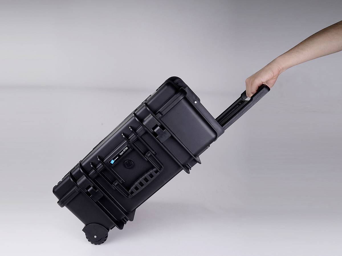 A hand pulls a black wheeled hard-shell suitcase with an extended handle across the floor.