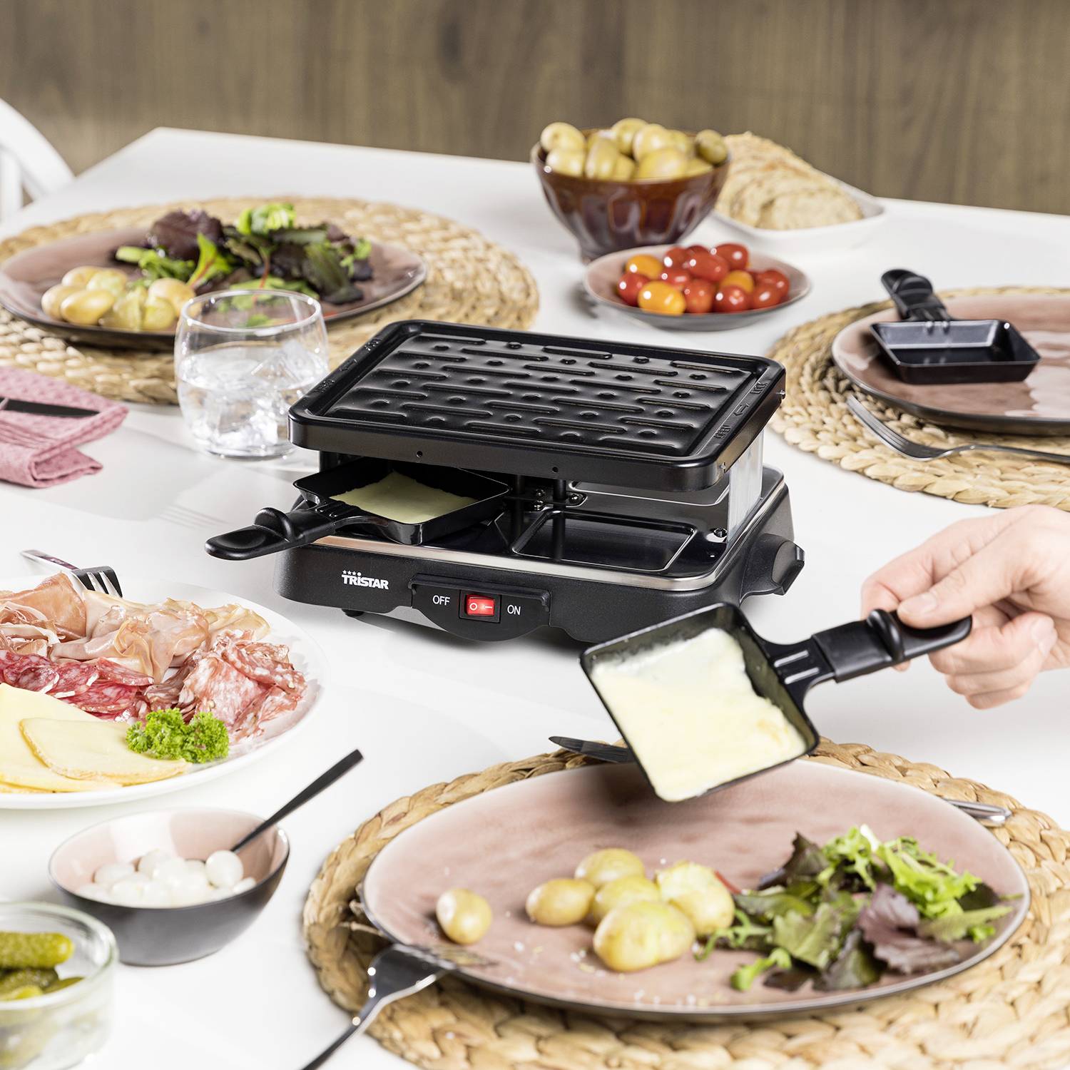 Raclette grill on a laid table with salad, potatoes, ham and tomatoes. A hand is swirling melted cheese onto the plate.