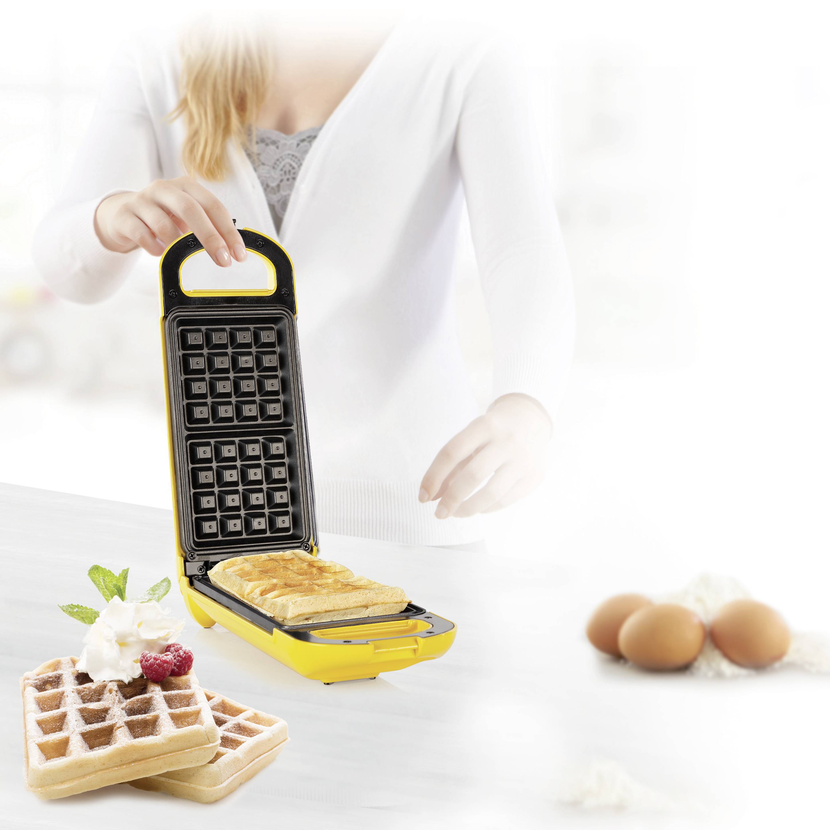 A person is preparing waffles using a waffle iron. On the table are raw eggs, flour, and a finished waffle with berries.