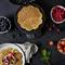 A waffle iron with freshly made waffles, surrounded by bowls of blueberries, raspberries and strawberries on a dark table.