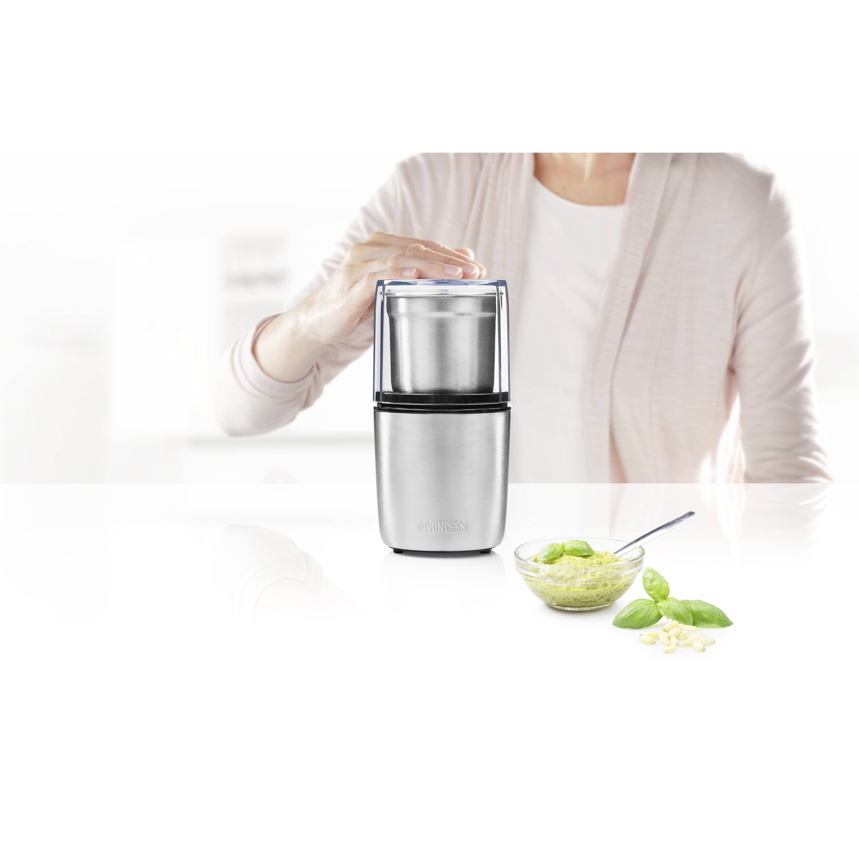 A person in a white top is using a coffee grinder. Next to it is a bowl of green pesto and basil leaves.
