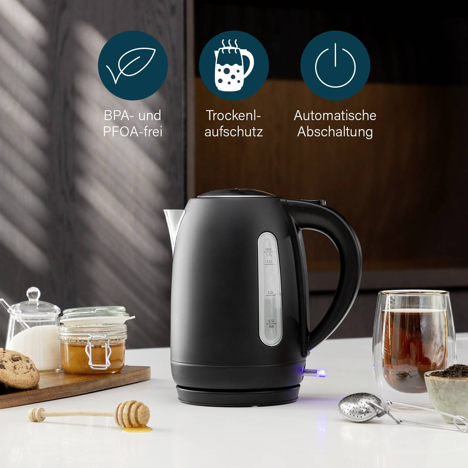 Black kettle on kitchen table, surrounded by glass with cutlery and food. Text: 'BPA and PFOA free', 'Boil-dry protection', 'Automatic switch-off'.