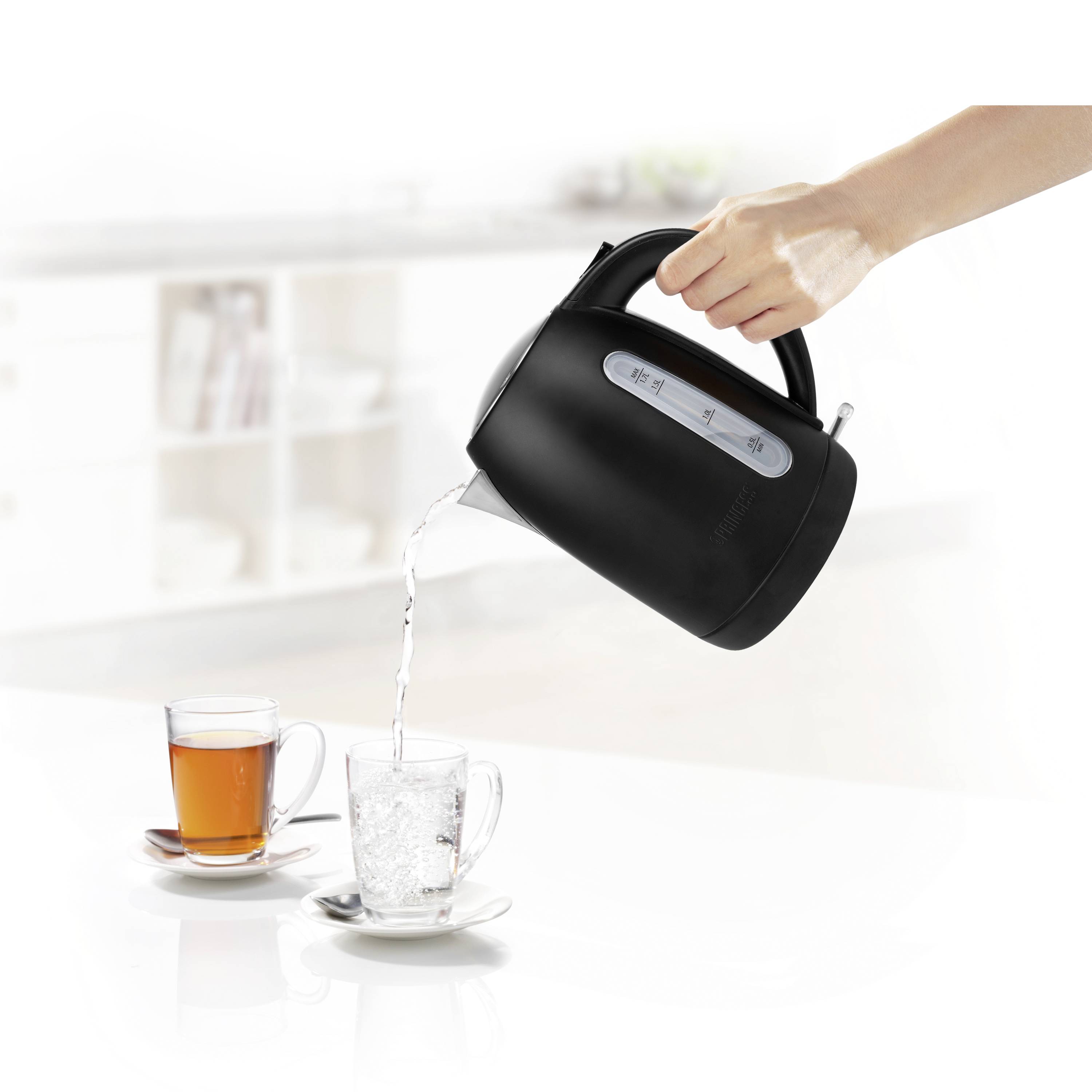 A hand is pouring hot water from a black kettle into a cup; beside it a cup of tea, both on saucers.