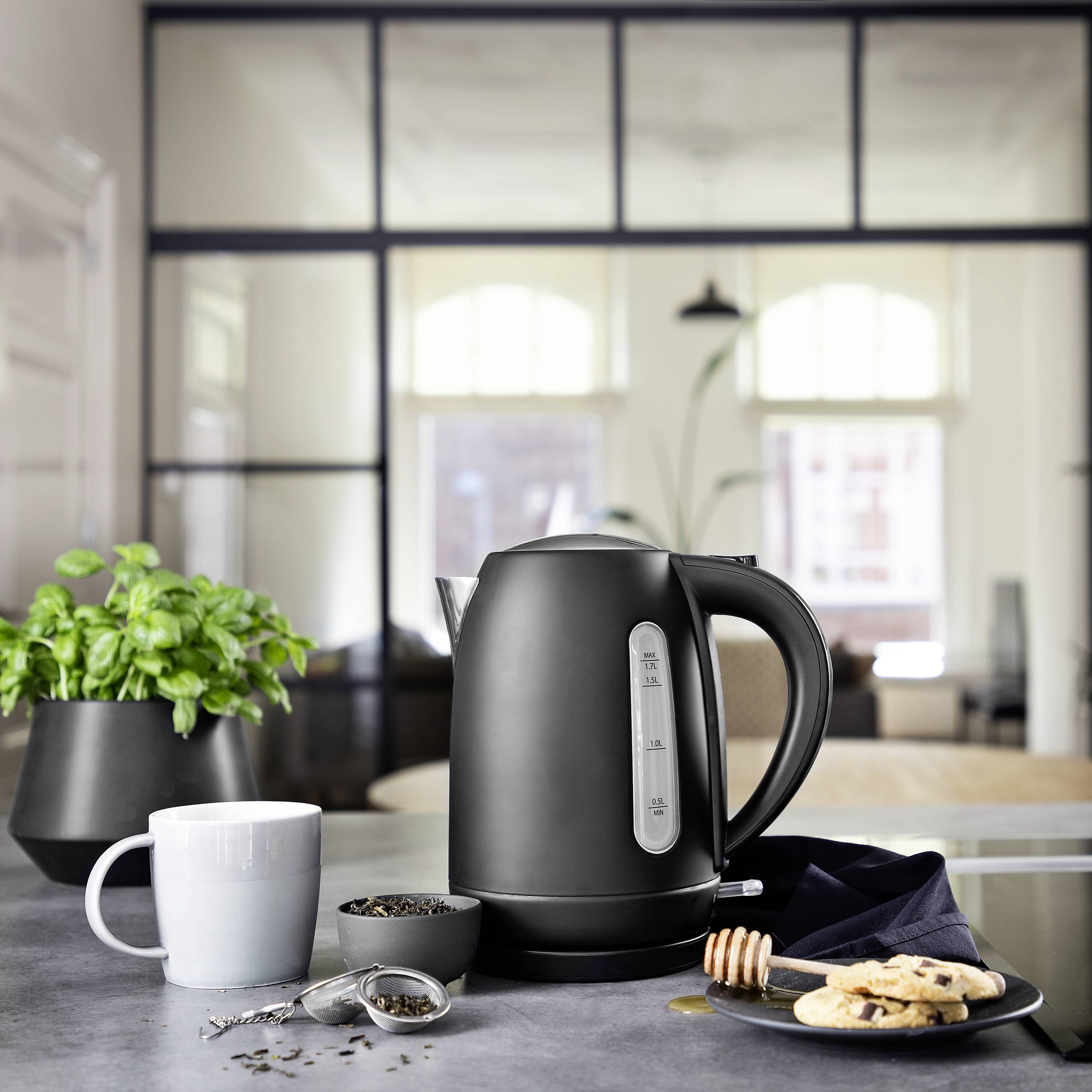 Black kettle on kitchen worktop, surrounded by a cup, loose tea, spoon and biscuits. Background with large window slightly out of focus.