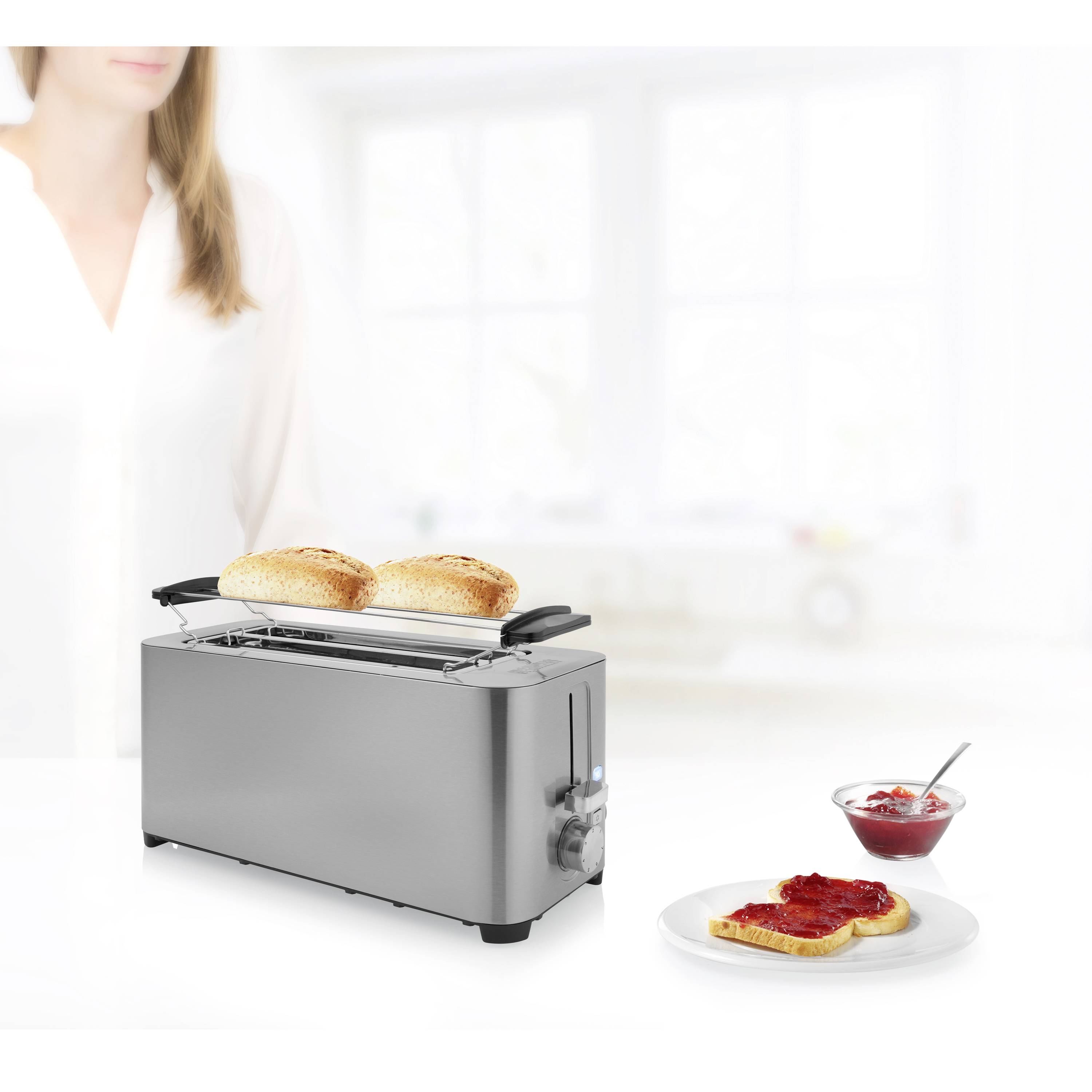 A silver two-slice toaster with two pieces of toast beside a plate of toast and red jam. A blurred person is visible in the background.