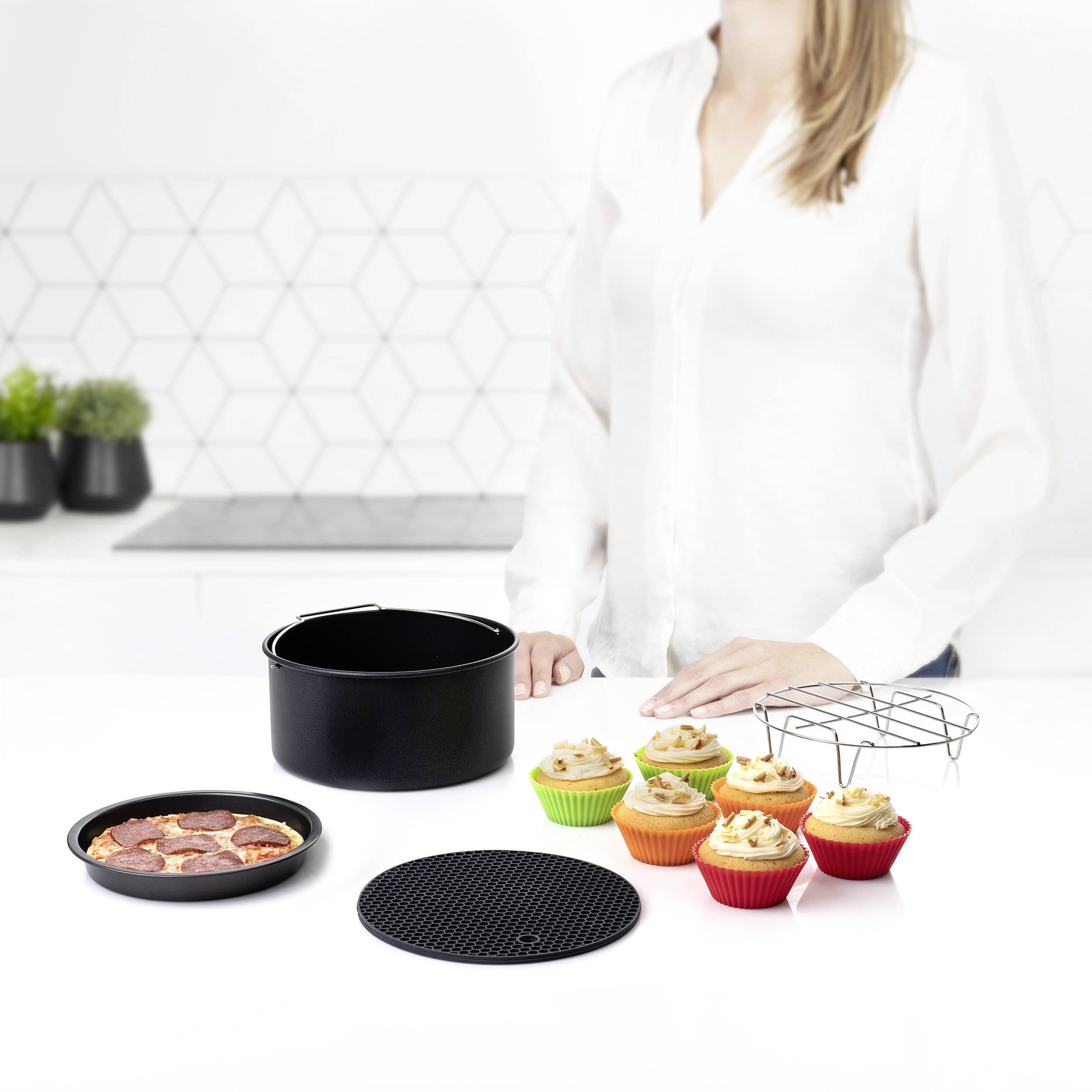 A person is standing in a modern kitchen. On the worktop, there is a cake, cupcakes in colourful cases, a cooling rack, and a baking tin.