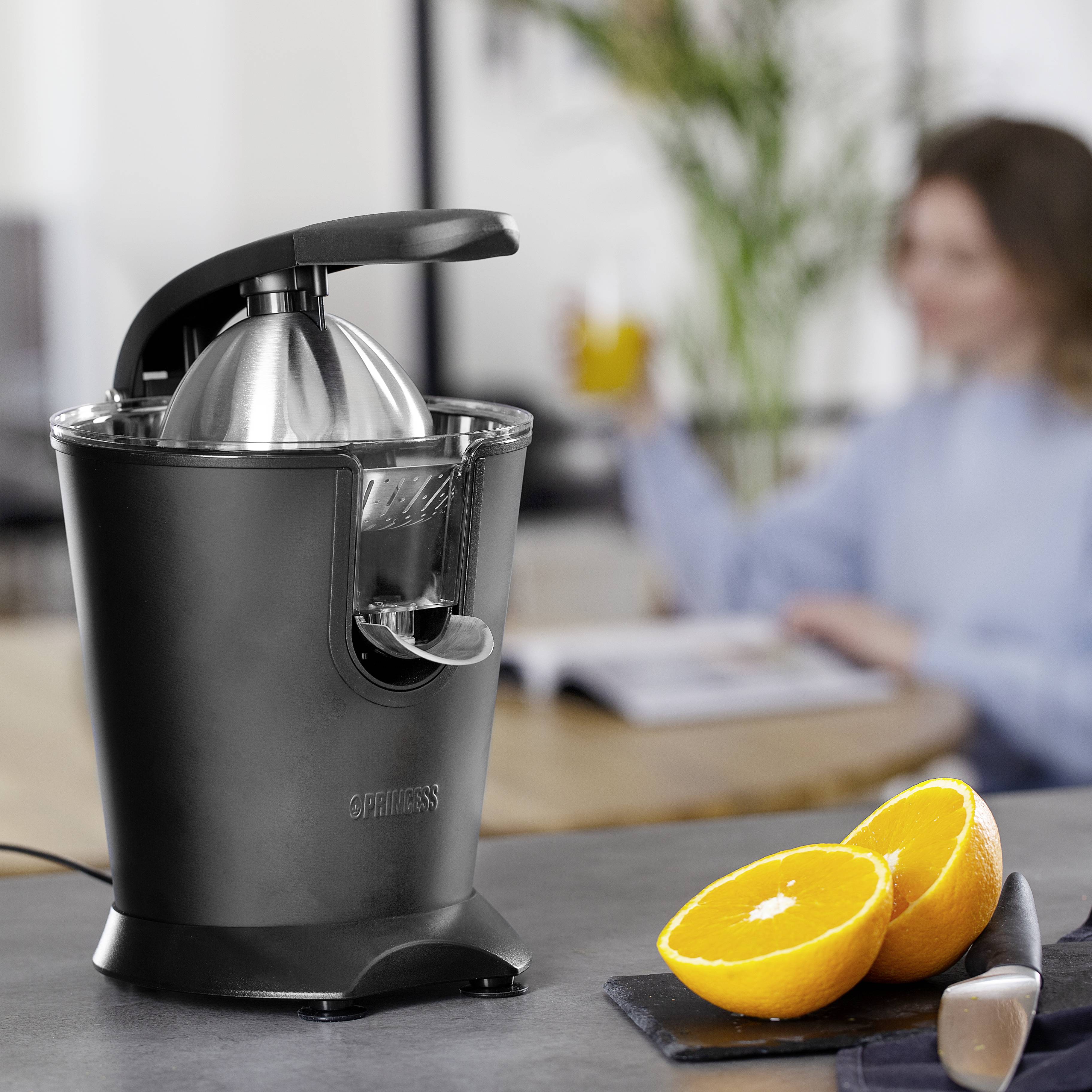 Electric juicer on a table, with halved oranges beside it. In the background, a blurry person holding a glass of juice.