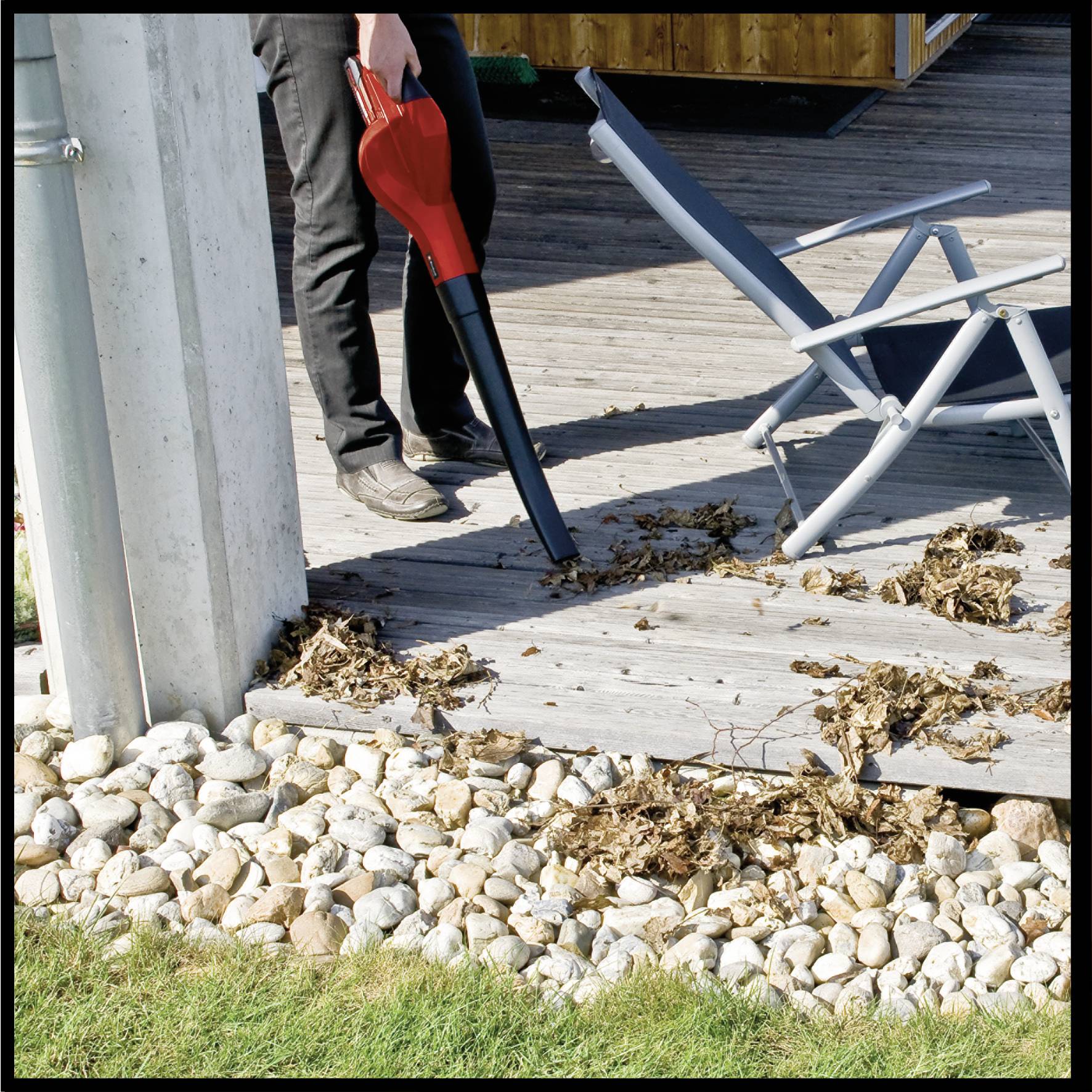 A person is removing leaves from a wooden decking using a leaf blower. A blue lounger is standing nearby.