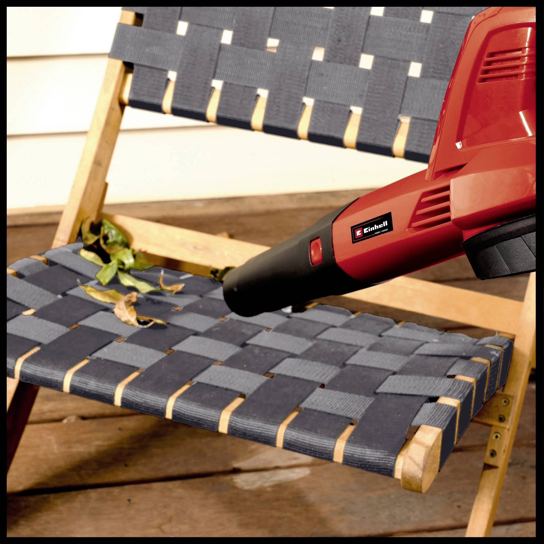 A red leaf blower is resting on a wooden chair with a blue, woven seat. Leaves are scattered across the chair.