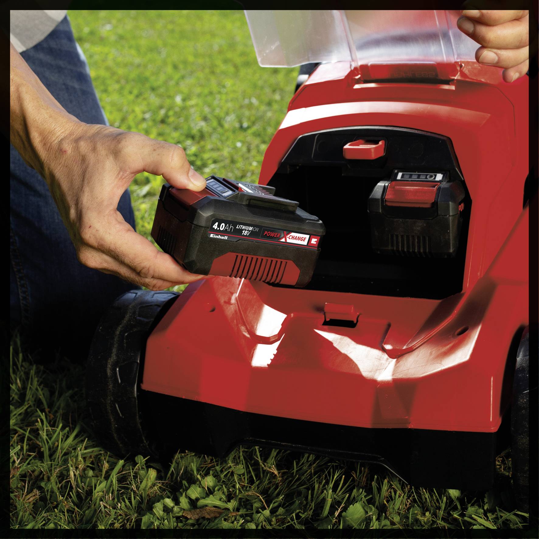 A person is inserting a battery into the battery compartment of a red lawnmower. The lawnmower is standing on a lawn outdoors.
