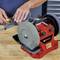 A person is grinding a tool with a red and black grinding device on a workbench. Hands are pressing the tool against the grinding wheel.