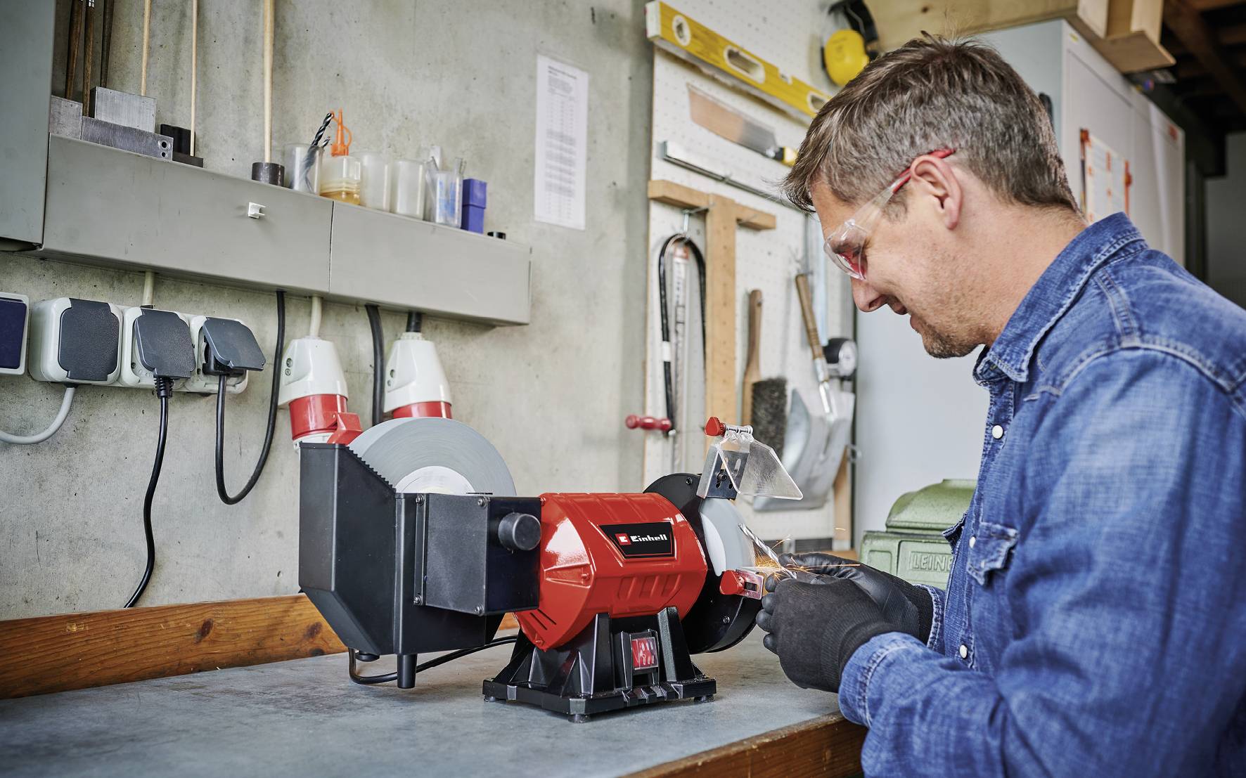 A person is grinding a tool on a grinding wheel in a workshop. Wearing safety glasses and gloves for protection.