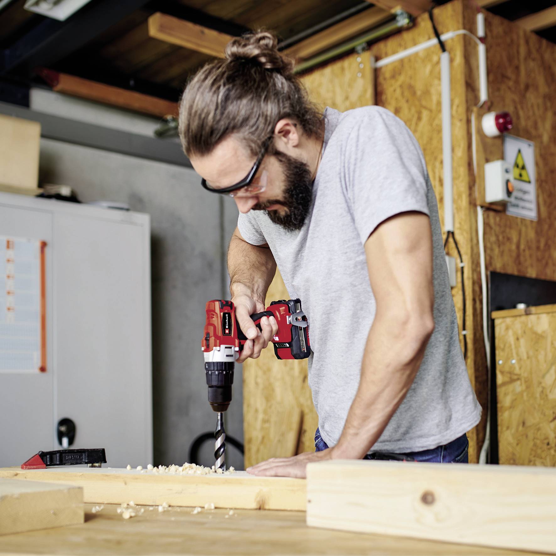 A man wearing glasses and a beard is drilling into a piece of wood with a red drill in a workshop setting.