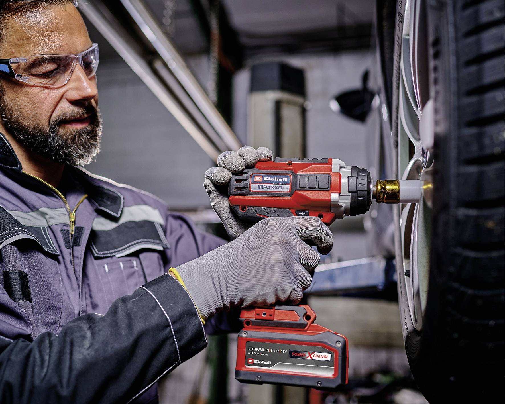 A mechanic in safety gear uses a power drill to tighten a tire on a vehicle's wheel in a garage setting.