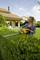 Woman wearing safety glasses trims hedge with electric hedge trimmer in garden. House with red tiles in the background.